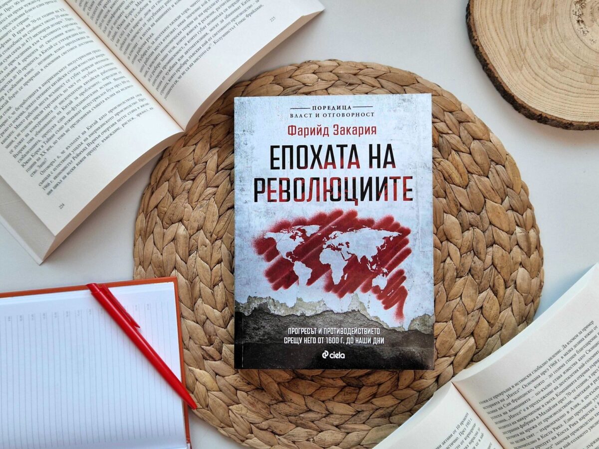 Корица на книгата „Епохата на революциите“ от Фарийд Закария, издание на „Сиела“, поставена върху плетена подложка, с отворени книги и тефтер наоколо.