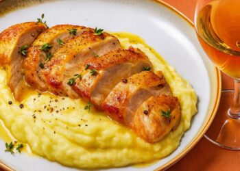 Апетитно сервирани запечени пилешки гърди върху кремообразно картофено пюре, гарнирани с пресни подправки и поднесени с чаша бяло вино.