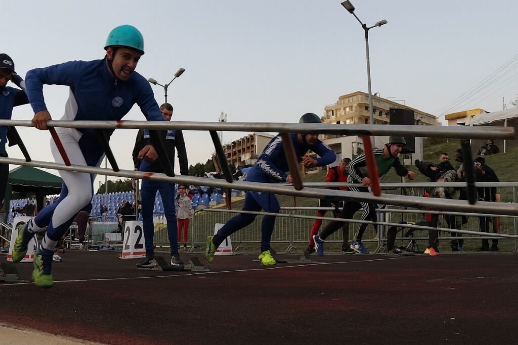 Състезатели в турнир по пожароприложен спорт прескачат препятствия на стадион, облечени в екипировка и каски.