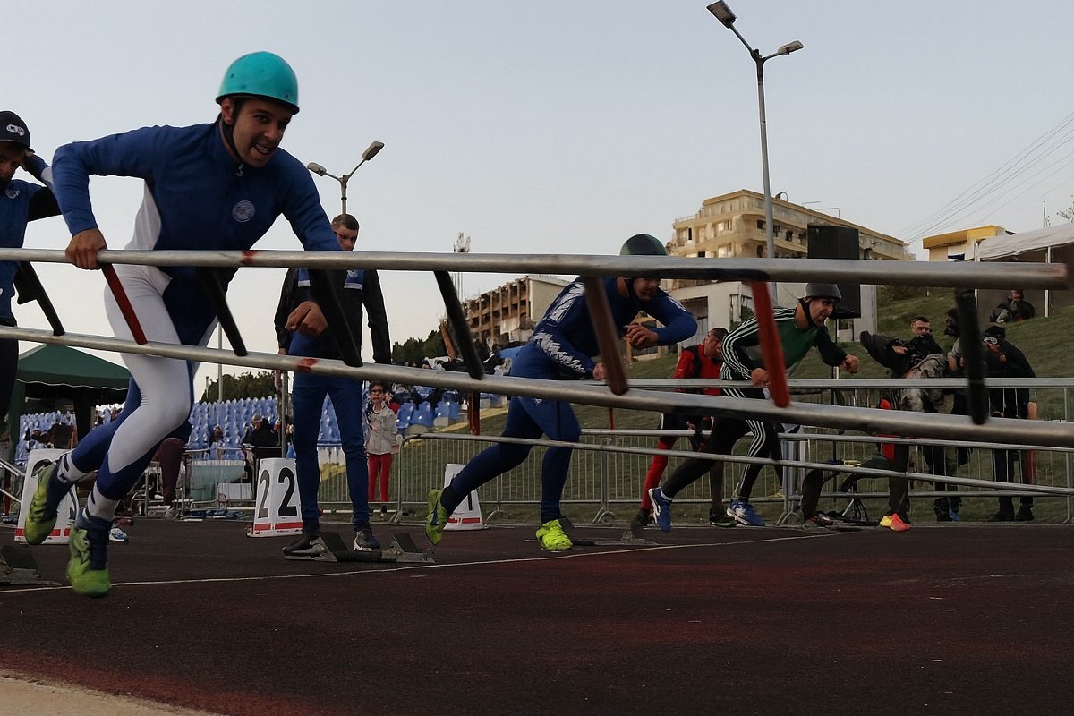 Състезатели в турнир по пожароприложен спорт прескачат препятствия на стадион, облечени в екипировка и каски.