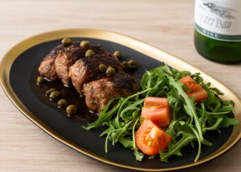 Елегантно сервирани свински медальони с балсамов сос и каперси, поднесени с рукола и домати върху черна чиния със златен ръб, в компания на бутилка бяло вино