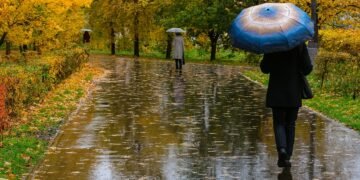 „Хора с чадъри вървят по мокра есенна алея след дъжд, сред жълти листа и дървета — типичен дъждовен ден по Черноморието.“