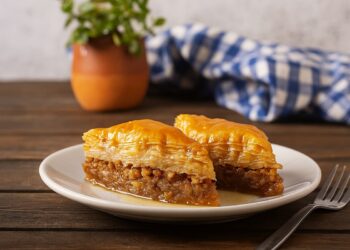 Традиционна гръцка баклава, поднесена в керамична чиния с орнаменти, на фон с бяло-сини гръцки мотиви и морска светлина, гарнирана с орехи и мед.