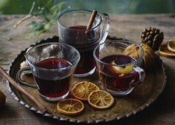 Три стъклени чаши с греяно вино, украсени с канелени пръчици и резени портокал, подредени върху дървена маса с коледен декор и топла, уютна светлина.