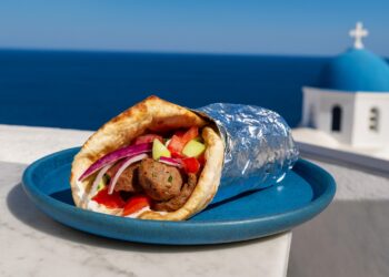 Гръцки гирос с агнешко, свежи зеленчуци и чеснов сос, увит в топла пита и алуминиево фолио, поднесен на синя чиния с гледка към морето в Гърция.