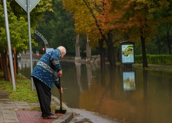 Възрастен мъж почиства шахта на наводнена улица през есента, за да може водата да се оттече.
