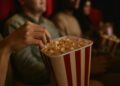 „Зрители в киносалон държат кофи с пуканки, докато гледат филм. Снимката е с топла светлина и уютна атмосфера, подходяща за статия за кино и филми.“