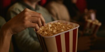 „Зрители в киносалон държат кофи с пуканки, докато гледат филм. Снимката е с топла светлина и уютна атмосфера, подходяща за статия за кино и филми.“