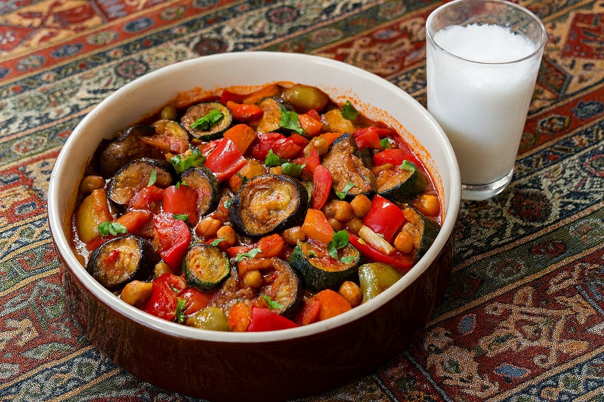 Ароматно ястие от печени зеленчуци и нахут в ориенталски стил, поднесено в глинен съд с чаша традиционна напитка върху шарена покривка.