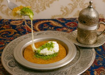 радиционно турско кюнефе, поднесено в сребърен съд с топящо се сирене и поръска от шамфъстъци, върху ориенталска покривка до чаша турско кафе в гравирана метална поставка.