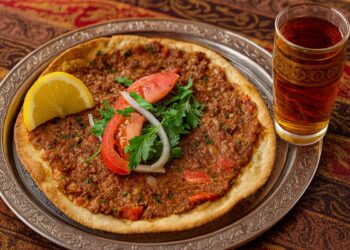 Турски лахмаджун – тънка ориенталска пица с мляно месо, домати, лук и магданоз, поднесена в ориенталски стил върху дървена дъска, гарнирана с лимон и свежа салата. До нея има традиционна чаша айрян, създаваща усещане за автентична турска кухня и домашна атмосфера.