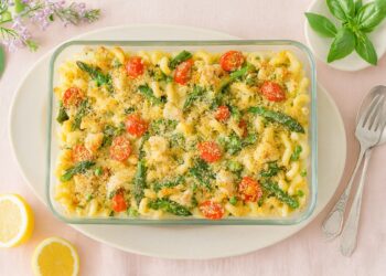 Печена паста Примавера с пиле, аспержи, грах, спанак и чери домати, поднесена в стъклена тавичка върху светлорозов плат и декорирана с лимони и свеж босилек