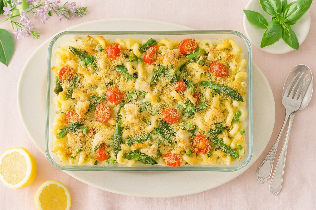 Печена паста Примавера с пиле, аспержи, грах, спанак и чери домати, поднесена в стъклена тавичка върху светлорозов плат и декорирана с лимони и свеж босилек