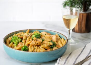 Чиния с паста пене с пиле Алфредо, броколи и сметаново-пармезанов сос, поднесена в синя керамична купа на мраморна повърхност, до чаша бяло вино и ленена салфетка.