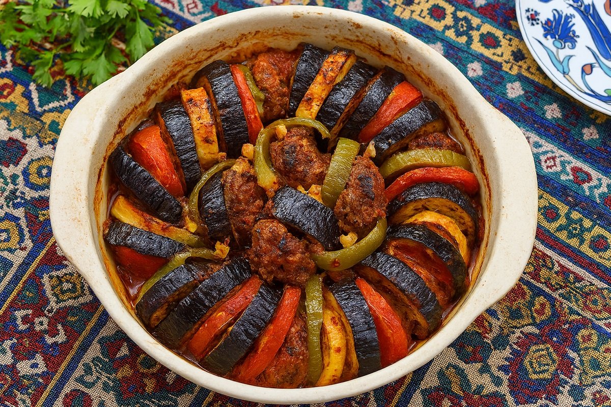 Печен патладжанен кебап с кюфтета, подреден в кръгла керамична тава, гарниран с домати, зелени чушки и ароматен сос, върху традиционна турска покривка.