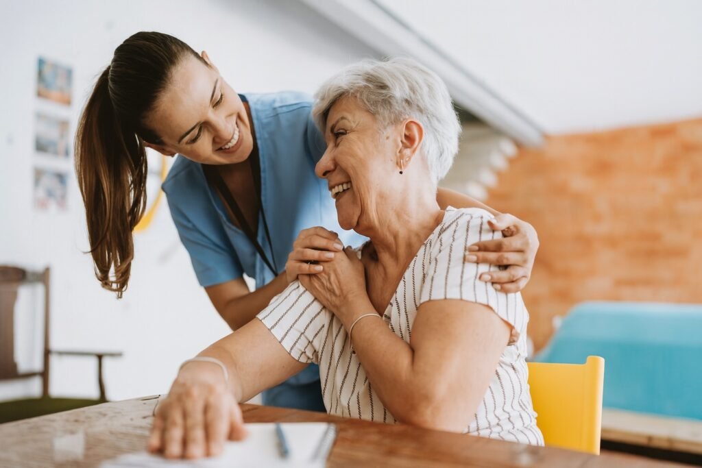 Млада медицинска сестра с усмивка подкрепя възрастна жена, като я докосва по рамото. И двете се смеят и излъчват топлина и доверие в уютна среда
