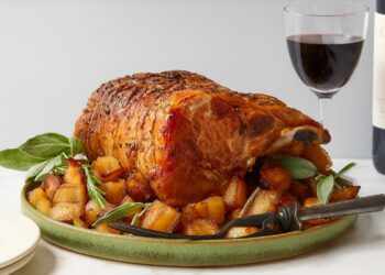 Празнично печено свинско каре с кост, гарнирано със златисти картофи и пресен розмарин, сервирано в зелена керамична чиния с чаша червено вино на заден план.