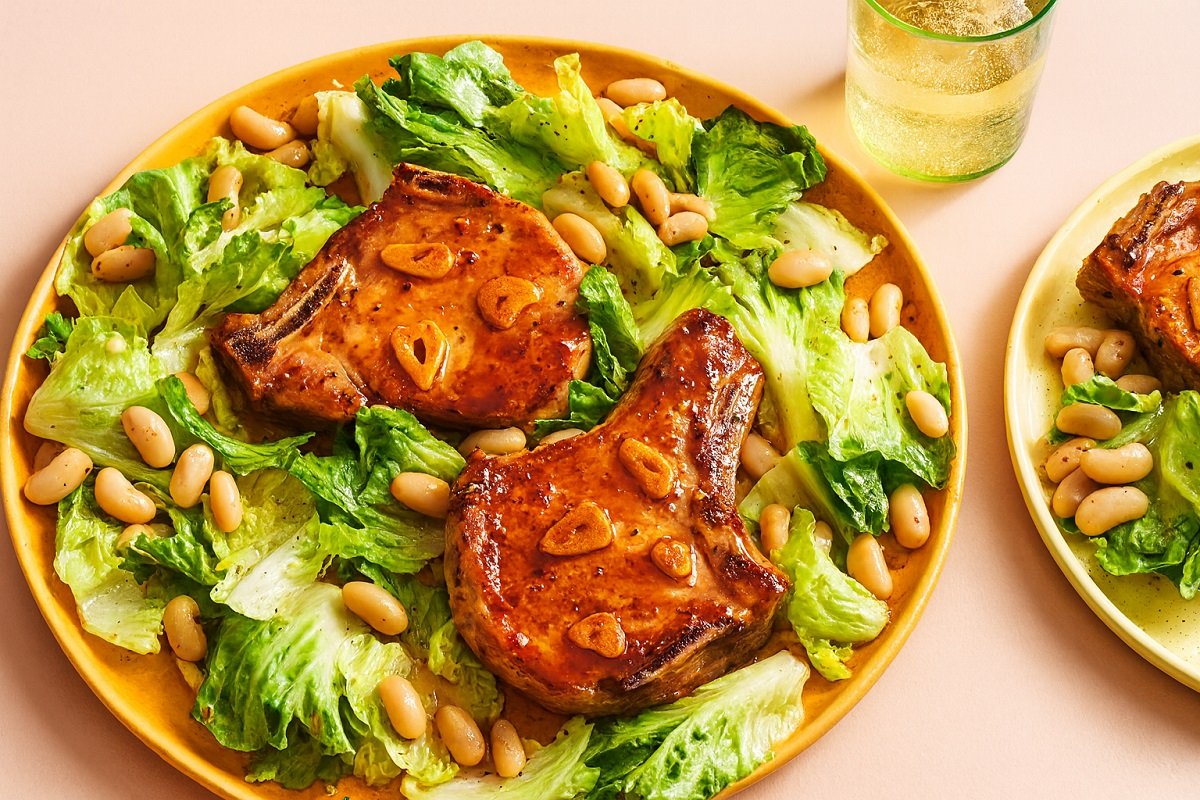 „Апетитни свински котлети с меден сос, поднесени с топла салата от бял боб и зелени листа върху елегантна керамична чиния, в уютна обстановка, подходяща за домашна вечеря.“