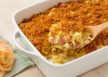 Ароматна запеканка с пилешко, шунка и разтопен кашкавал, изпечена до златисто в керамична тава, поднесена на дървена маса с цветя и чаша бяло вино на заден план.