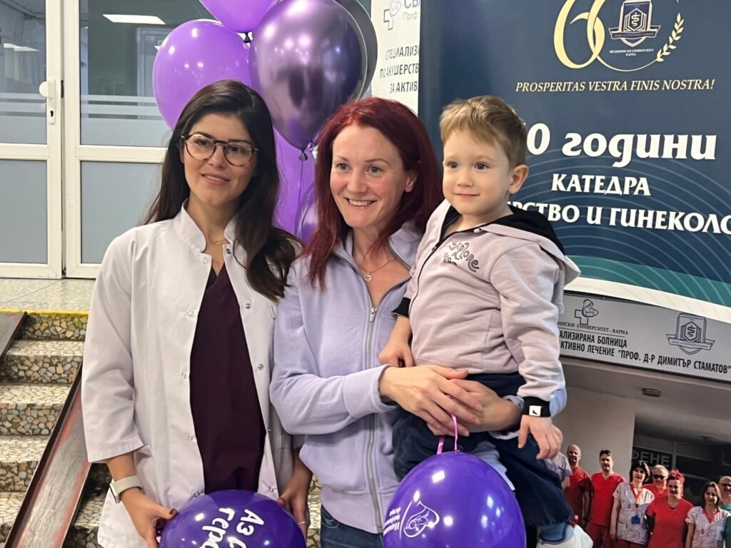 Медицински специалист и майка с малко момче позират усмихнати по повод събитие за недоносените деца в АГ болница Варна, с лилави балони и информационни табла на заден план.