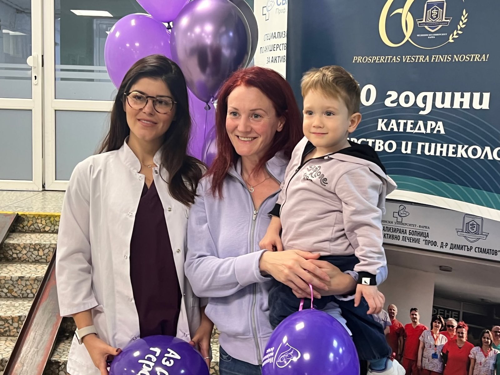 Медицински специалист и майка с малко момче позират усмихнати по повод събитие за недоносените деца в АГ болница Варна, с лилави балони и информационни табла на заден план.