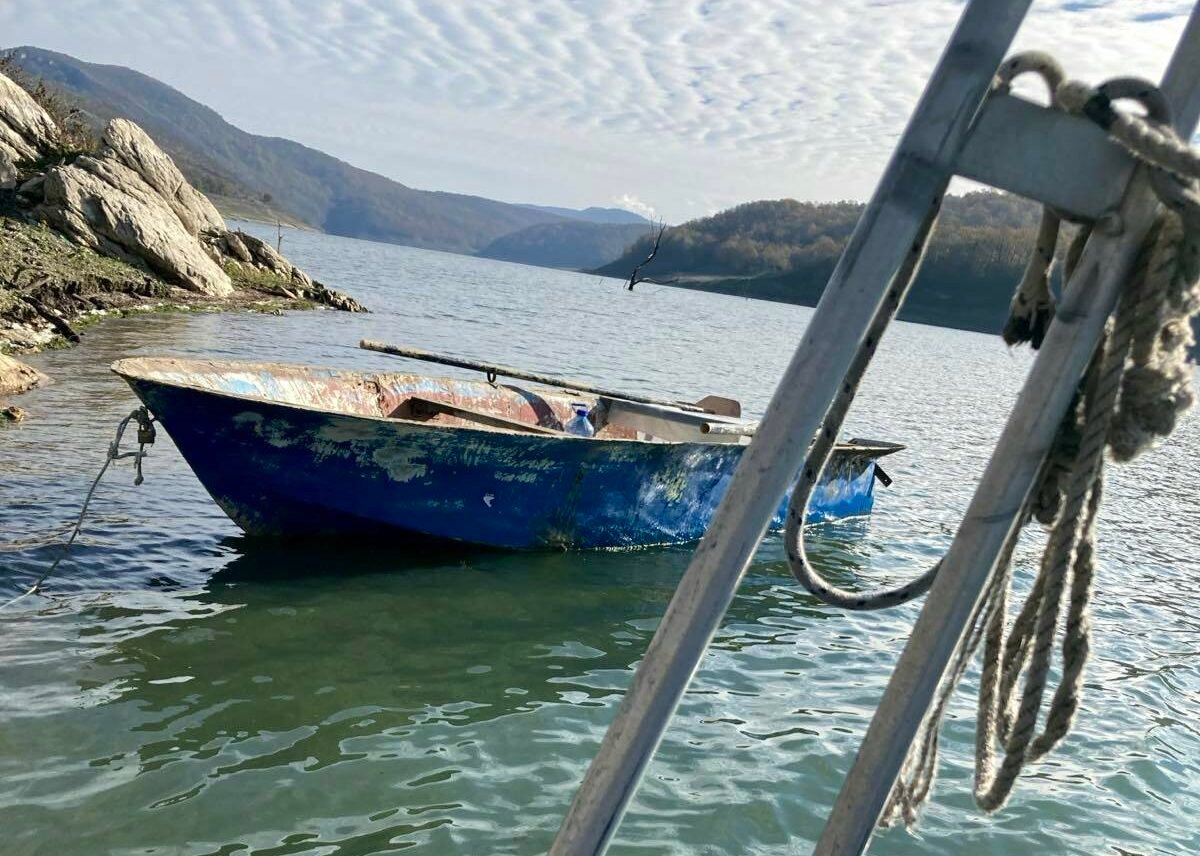 Синя метална лодка е привързана край скалист бряг в язовир „Цонево“, със спокойна вода и планински хълмове на заден план под облачно небе.