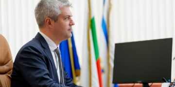 Профил на Благомир Коцев, седнал в кабинета си пред компютър, с национални и общински знамена на заден план.