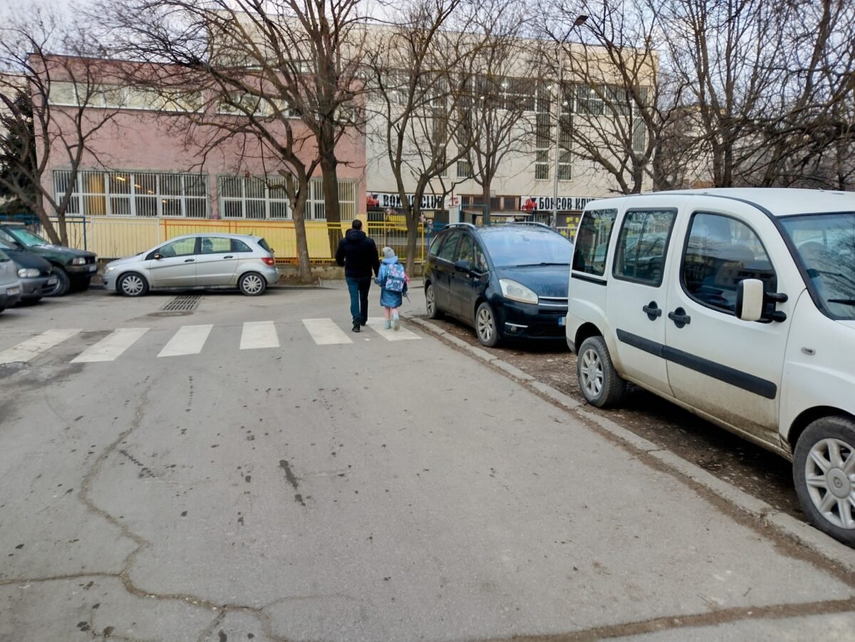 Баща и дете с ученическа раница вървят по улица до училище, а от двете страни на пътя са паркирани автомобили, някои върху тротоара.