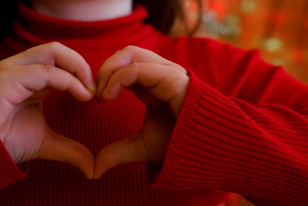 Две детски ръце оформят сърце върху червен пуловер на меко замъглен празничен фон.