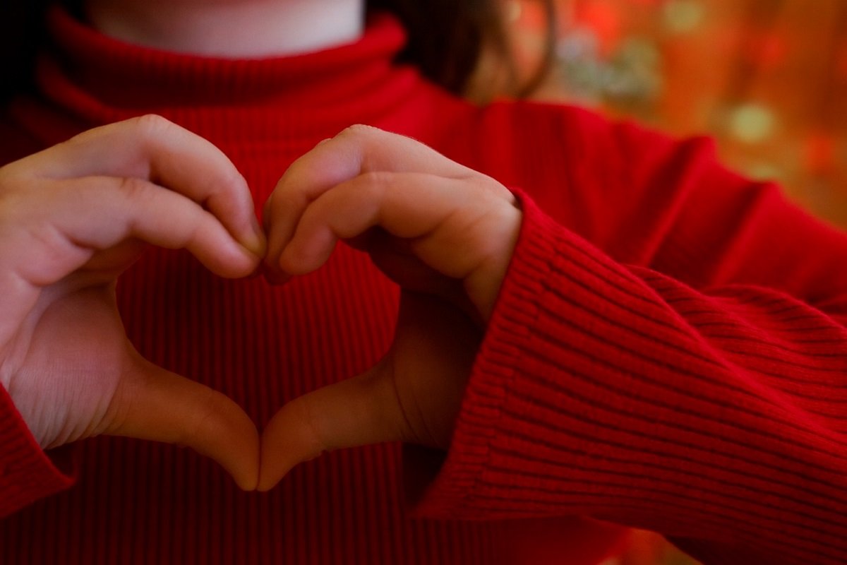 Две детски ръце оформят сърце върху червен пуловер на меко замъглен празничен фон.