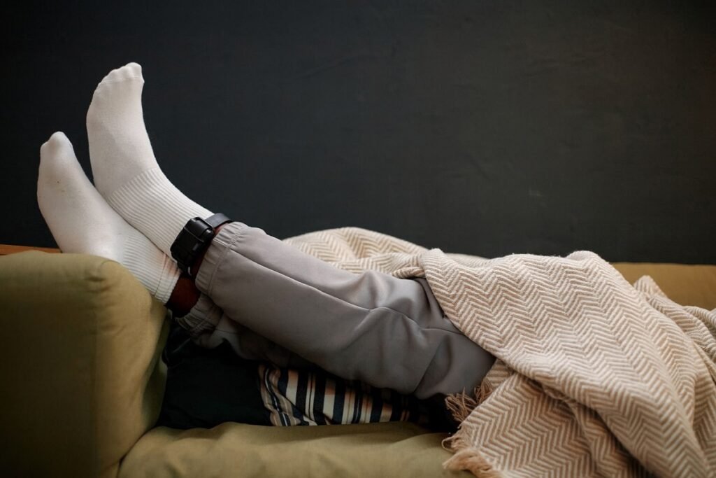 Крака на мъж, легнал на диван под одеяло, с поставена електронна гривна за домашен арест на глезена.