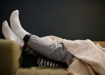 Крака на мъж, легнал на диван под одеяло, с поставена електронна гривна за домашен арест на глезена.