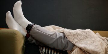 Крака на мъж, легнал на диван под одеяло, с поставена електронна гривна за домашен арест на глезена.