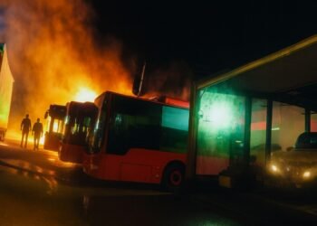 Пожар в автобусно депо през нощта с ярки пламъци и силно осветени автобуси, заснети отстрани.