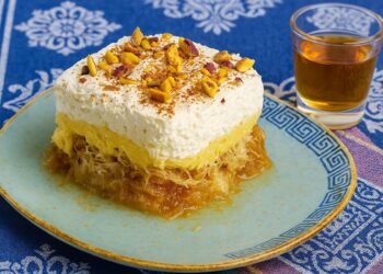 Парче гръцки десерт Екмек Кадаиф с крем, бита сметана и шамфъстък, поднесено върху чиния с гръцки орнаменти до чаша питие – автентичен средиземноморски вкус.