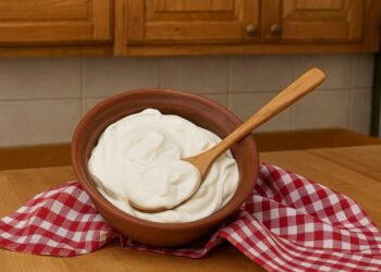 Купа с гъсто домашно гръцко кисело мляко и дървена лъжица върху дървена кухненска повърхност.