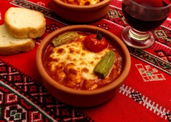 Традиционно българско ястие, поднесено в глинени купички с разтопен кашкавал, зеленчуци и доматен сос, върху дървена маса с български орнаменти и чаша червено вино до тях.