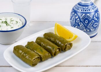 Традиционни гръцки лозови сарми (Dolmades) сервирани върху бяло плато с копър и лимон, до тях купичка с дзадзики и чаша узо на дървена маса с морски фон – типична средиземноморска сцена