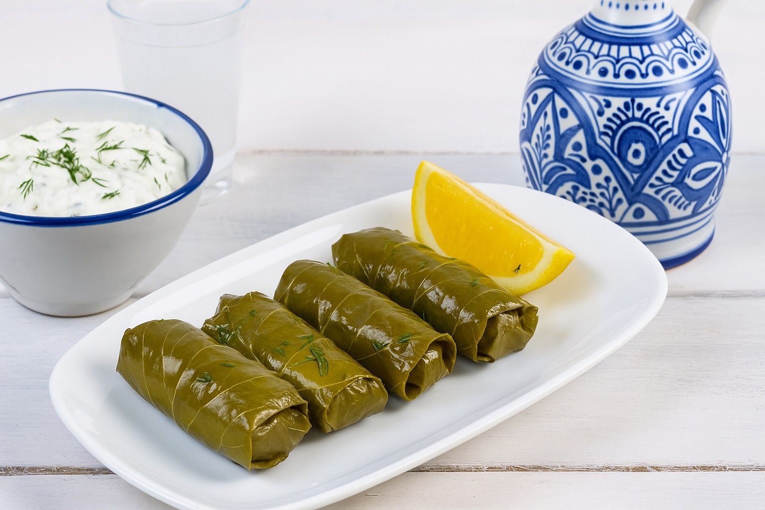 Традиционни гръцки лозови сарми (Dolmades) сервирани върху бяло плато с копър и лимон, до тях купичка с дзадзики и чаша узо на дървена маса с морски фон – типична средиземноморска сцена