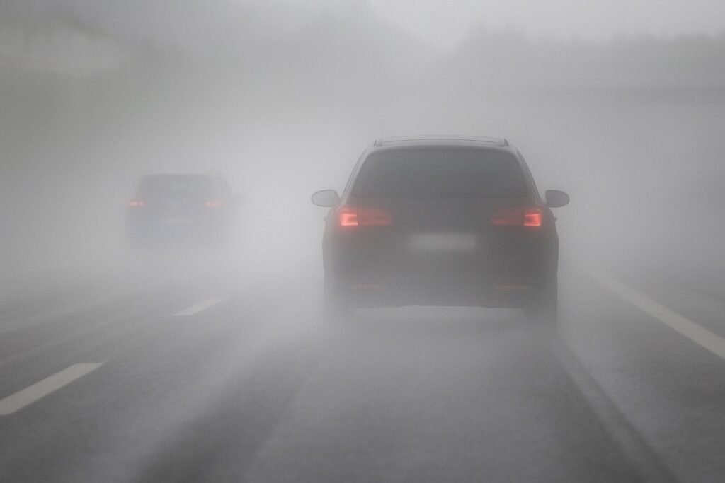 Две коли се движат по път, обгърнат от гъста мъгла, с ограничена видимост и влажна настилка, като червените им задни светлини проблясват в сивата атмосферна среда.