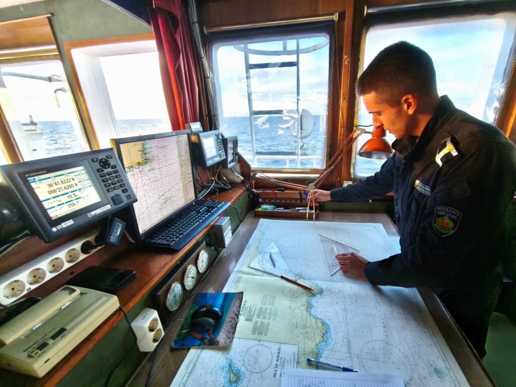 Млад курсант на борда на научноизследователския кораб „Св. св. Кирил и Методий“ работи върху навигационна карта на мостика, заобиколен от навигационни уреди и морски изглед през прозореца.