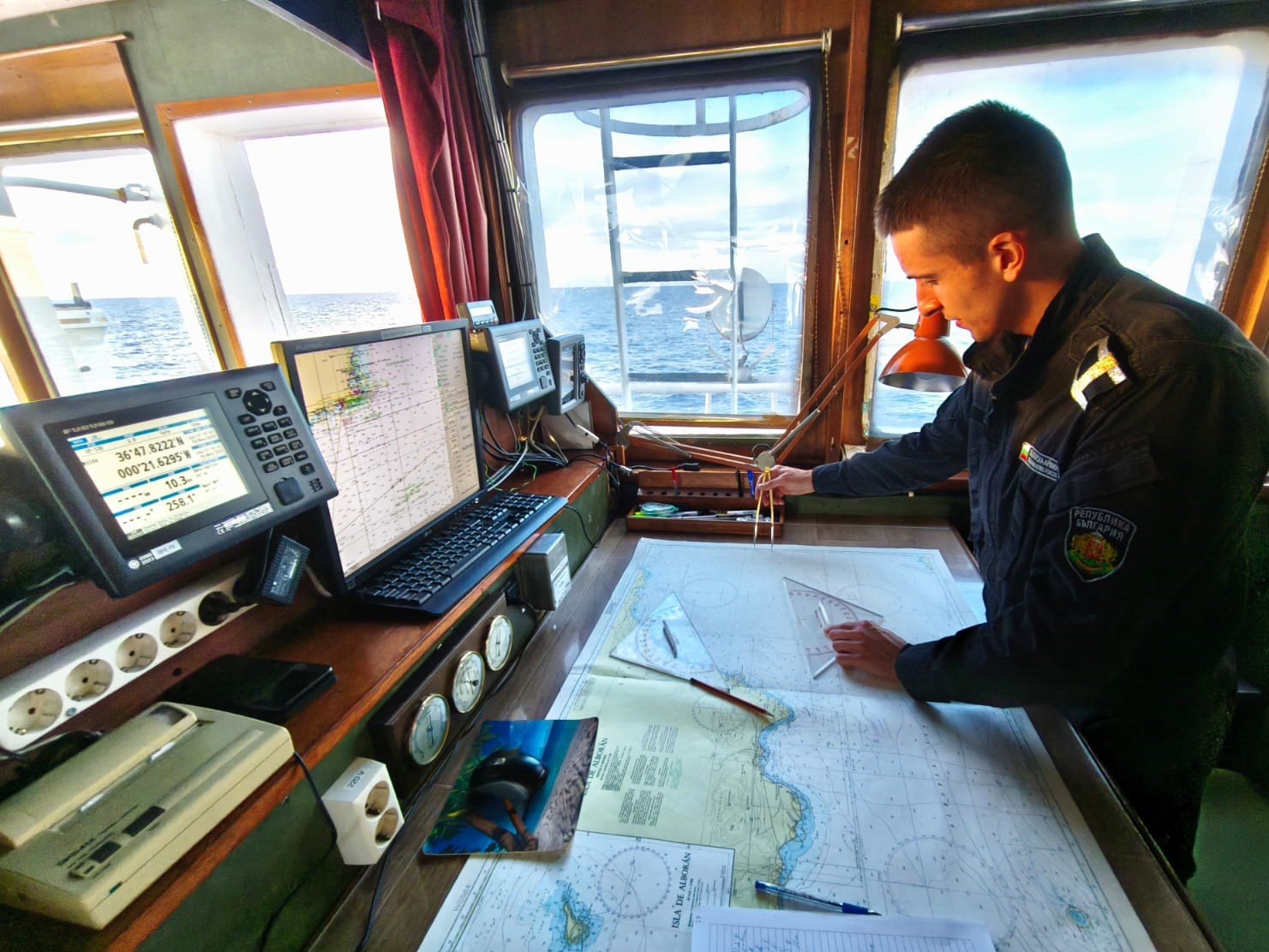 Млад курсант на борда на научноизследователския кораб „Св. св. Кирил и Методий“ работи върху навигационна карта на мостика, заобиколен от навигационни уреди и морски изглед през прозореца.