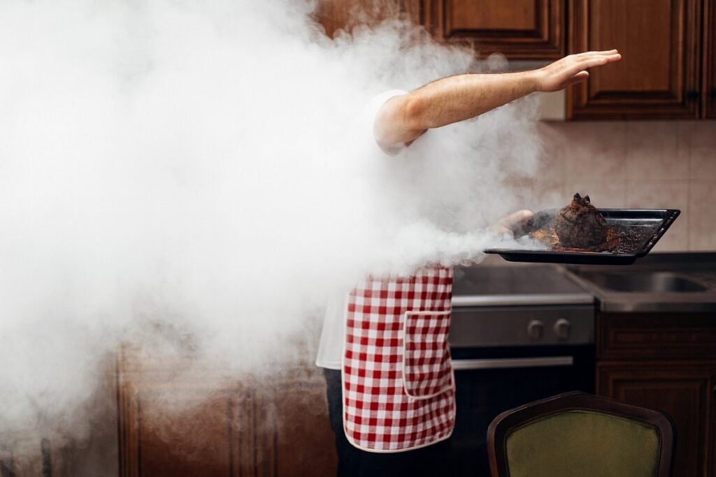 Голямо количество дим изпълва кухня, докато човек с престилка изнася силно изгоряла храна от фурната върху метална тава