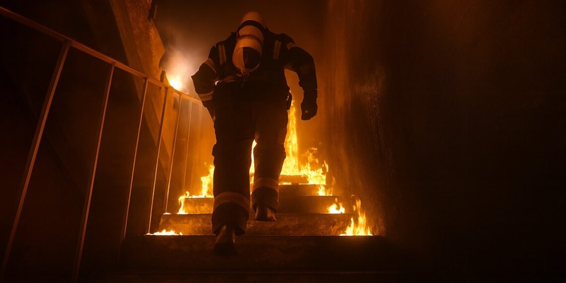 Пожарникар се изкачва по горящо стълбище в задимен коридор по време на инцидент, обграден от пламъци и осветен от оранжева светлина.