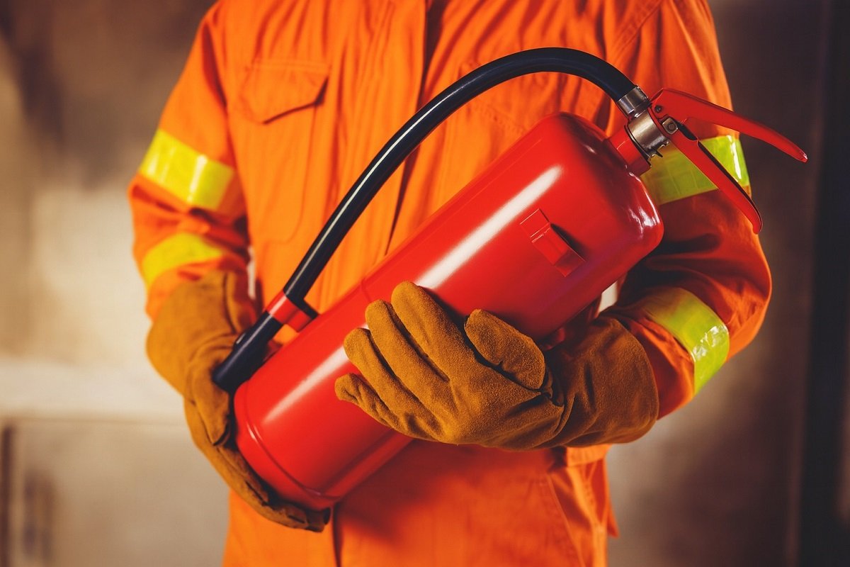 Пожарникар в защитно облекло държи червен пожарогасител с маркуч в ръце.