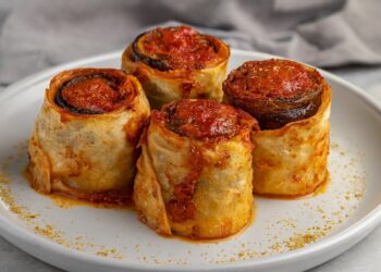 Хрупкави рулца с патладжан и кайма, увити в тънка кора и запечени с доматен сос, поднесени върху светла чиния в турско-битов стил, подходящи за сервиране.
