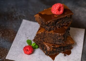 Шоколадено брауни, подредено на три слоя, полято с разтопен шоколад и декорирано с пресна малина и листо мента, заснето върху пергаментова хартия в хоризонтален формат.