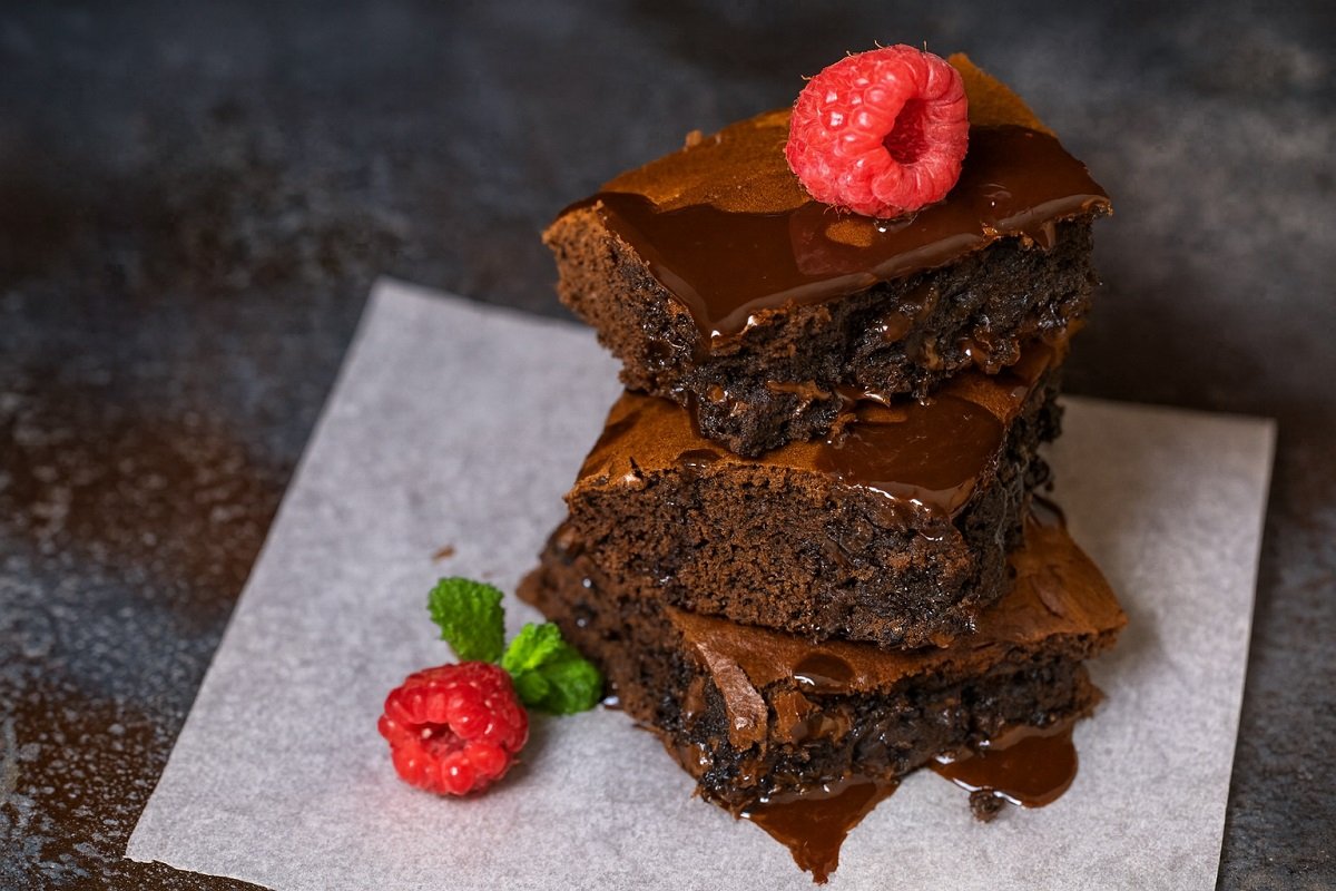 Шоколадено брауни, подредено на три слоя, полято с разтопен шоколад и декорирано с пресна малина и листо мента, заснето върху пергаментова хартия в хоризонтален формат.