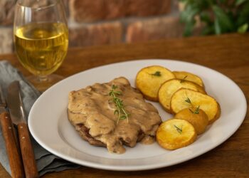 Домашно приготвен Стейк Даян с гъбен сметанов сос, сервиран с печени картофи и чаша бяло вино на дървена маса в уютна домашна обстановка.