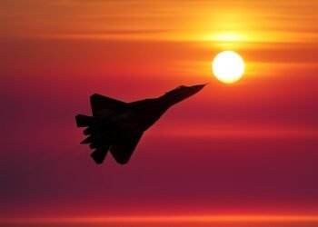 Силует на военен изтребител, летящ срещу ярък оранжево-червен залез с ниско стоящо слънце на хоризонта.